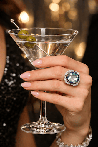 A close-up of a woman's hand holding a martini glass garnished with green olives, adorned with a stunning aquamarine and diamond ring, highlighting exquisite jewelry selection at KP Gems.